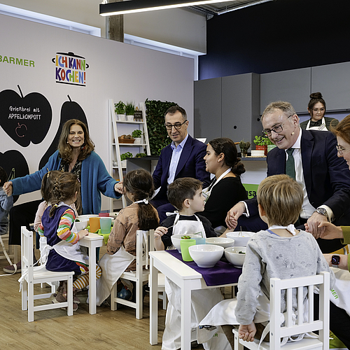 Foto von einer aufgebauten Küche in einem Studio mit Kindertischen und Kinderstühlen. An den Tischen sitzen Kinder und Erwachsene und reichen sich die Hände, um "Guten Appetit" zu sagen.